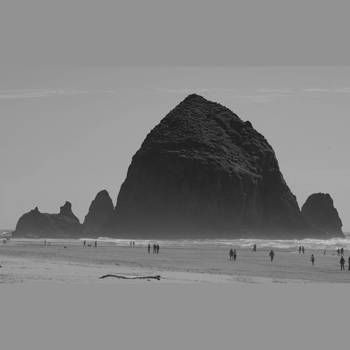 Black and white landscape photograph of Haystack Rock on a beach, with small silhouettes of people walking along the shoreline.