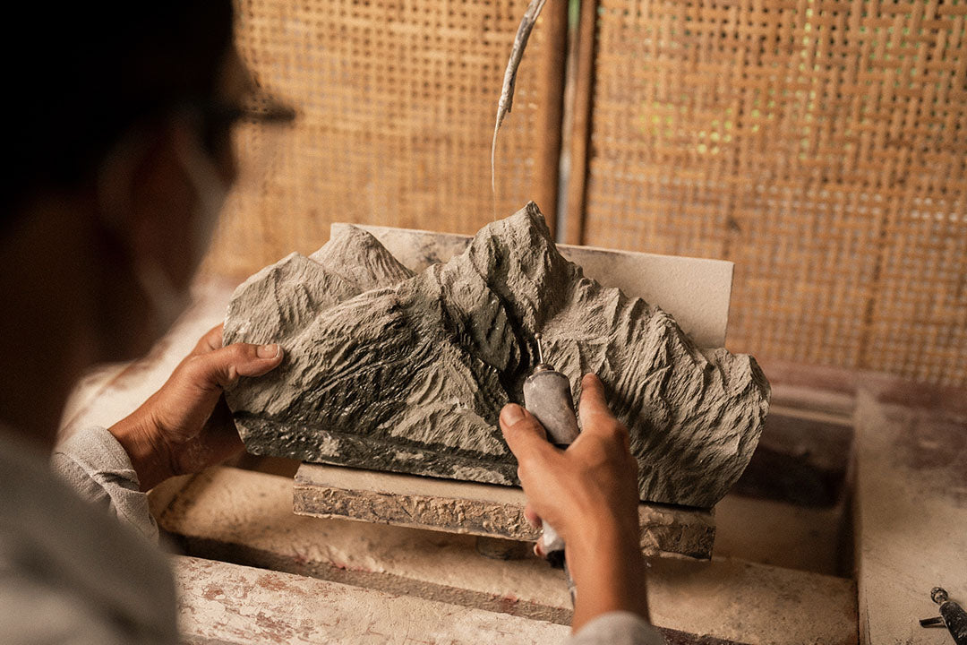 Artisan carving a medium Everest sculpture from dark gray stone, refining details with a handheld rotary tool in a workshop setting.