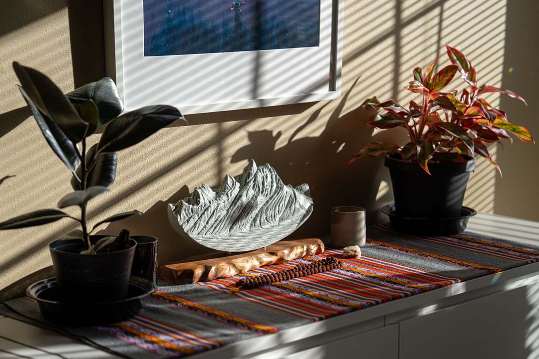 Medium Grand Teton sculpture in light gray stone on a rustic wood base, displayed on a decorative shelf with potted plants and woven fabric.