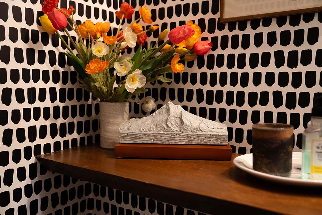 Small Mt. Hood sculpture in cream color stone on a mahogany copper wood base, placed on a wooden shelf beside a vase of flowers and a patterned wall